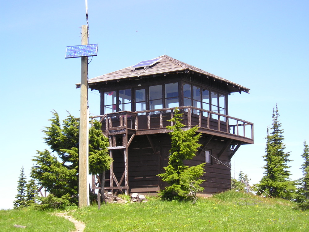 A small square building with large windows on all four sides and a wrap around porch on a grassy hilltop. 