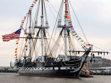 Three masted sailing ship floating in water. Her hull is painted a dark navy blue. Signal flags decorate her from the bowsprit up to the top of the foremast, over the main, down the mizzen and to the stern of the ship. A large american flag flies off the stern. A white strip runs down the middle broad side of her, highlighting the many cannon tubes protruding from her hull.