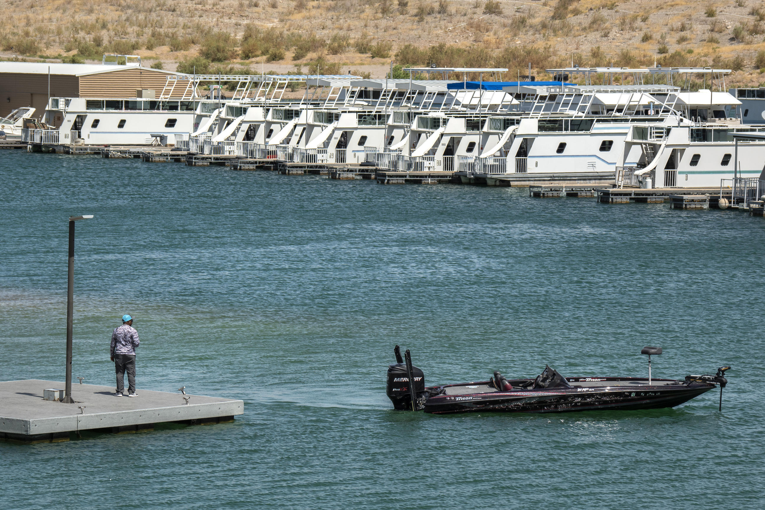 man stands on dock lower left, powerboat lower right, houseboats at marina in background