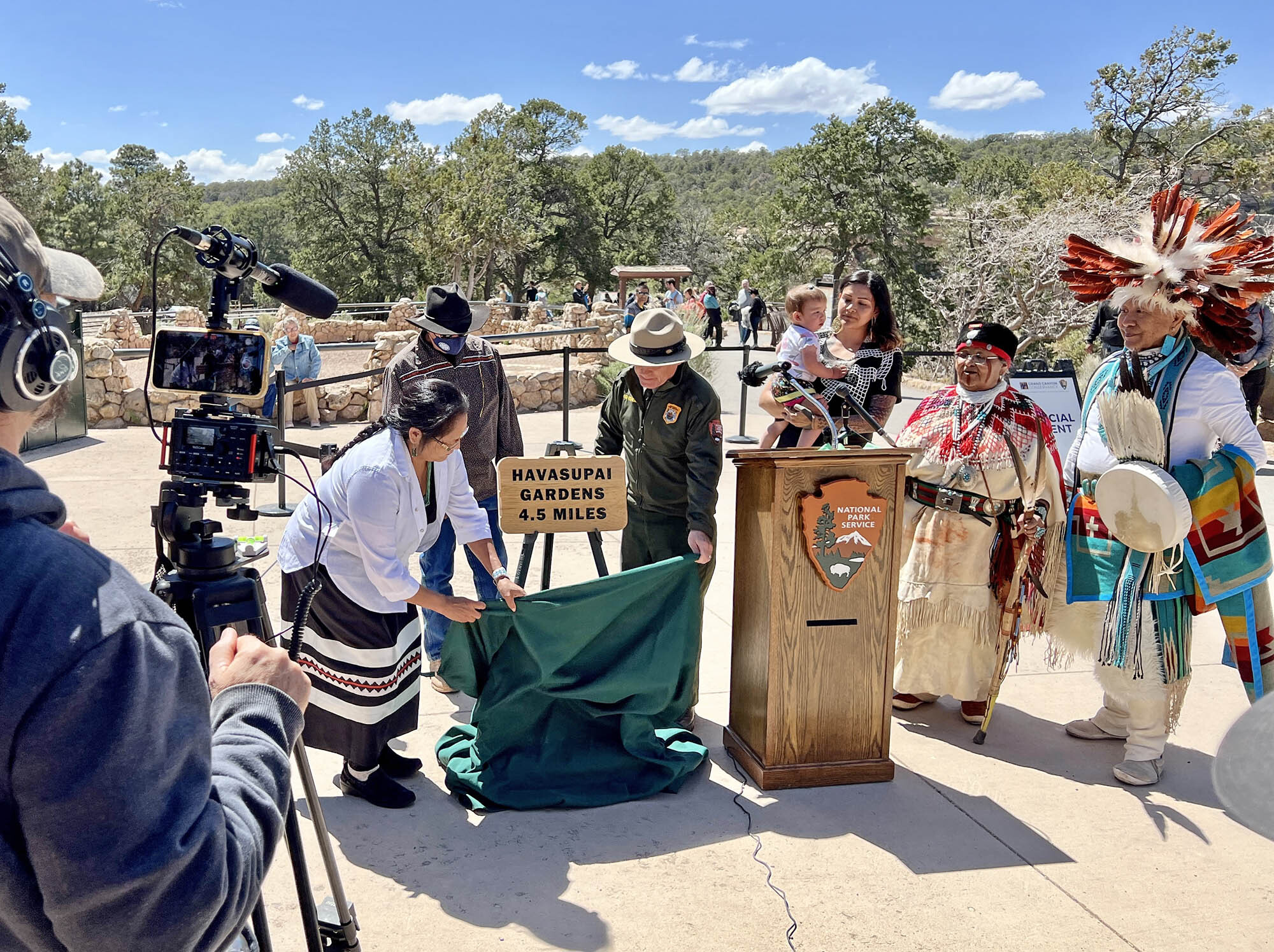 A woman and a man unveil a sign that reads "Havasupai Gardens 4.5 Miles" surround by people and a man with a camera man.