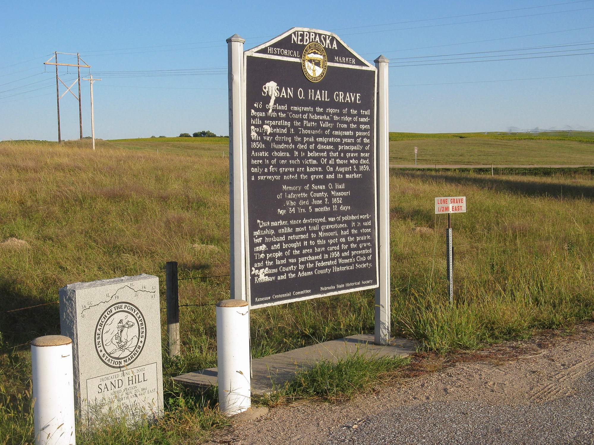 Large, blue metal sign with white colored text on Susan O. Hail Grave and a stone monument marking the Sand Hill Station Location