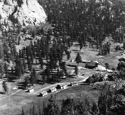 A row of four cabins along a road is seen from a distance, surrounded by pine trees and other structures.