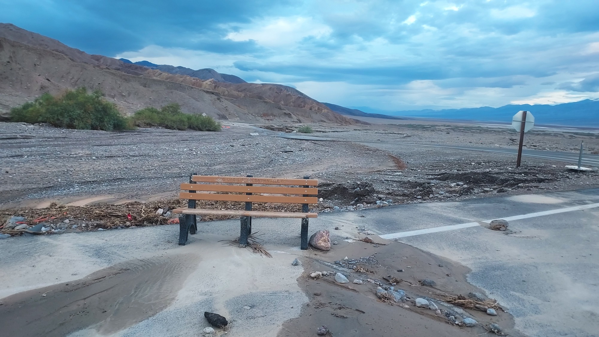 A brown wooden bench is misplaced on pavement with a cloudy mountain landscape behind it.