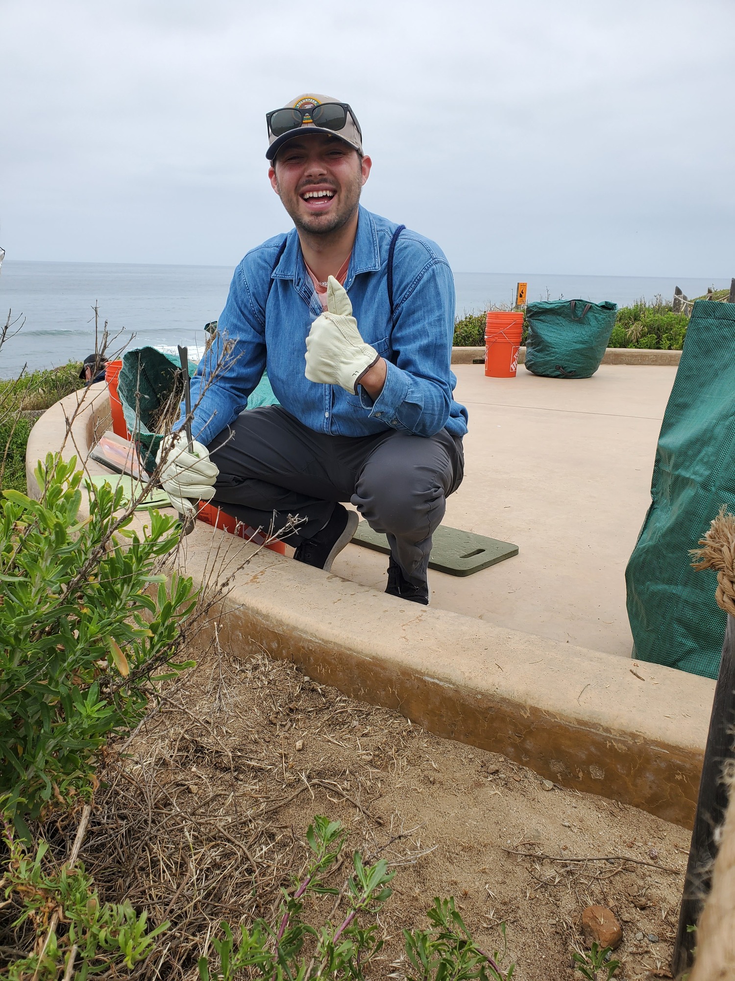 Weed Warriors (Habitat Restoration) volunteer gives a thumbs up as he kneels next to a newly restored segment of the Coastal Trail. Behind him is the gray Pacific Ocean and cloudy sky, along with the equipment of other volunteers. 