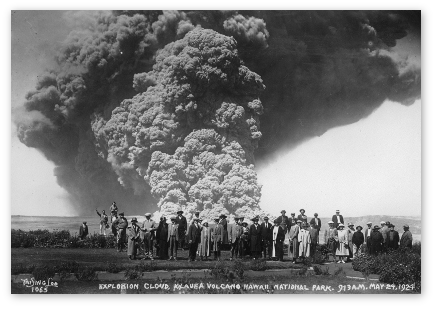 A 1924 black and white photo of a crowd standing at a volcanic overlook with a giant billowing cloud of ash and debris wafting behind them. 
