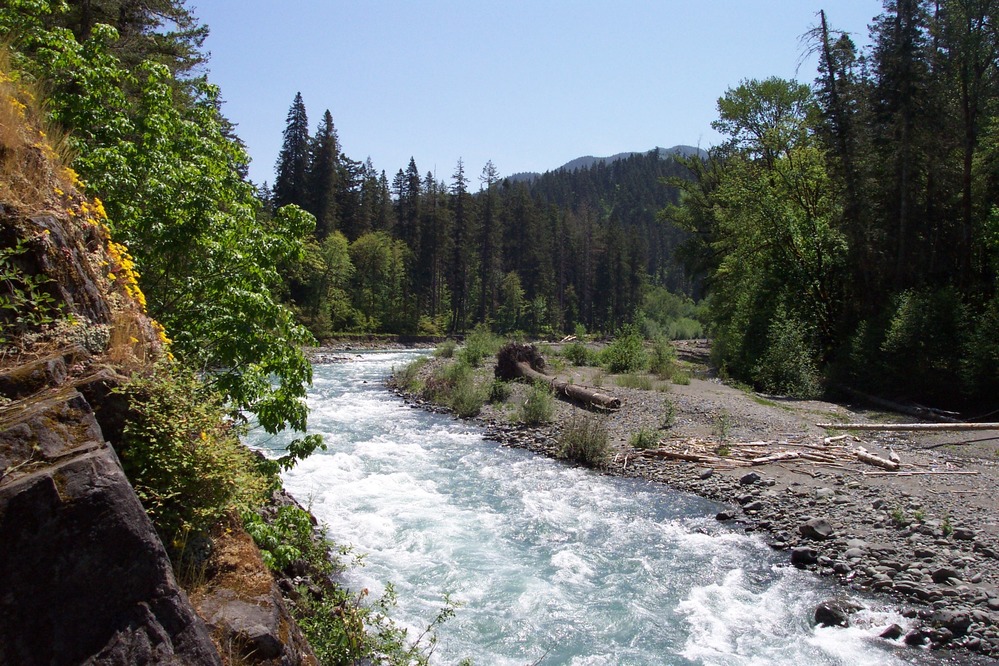 A rushing river flows between a cliffside and a rocky bank.