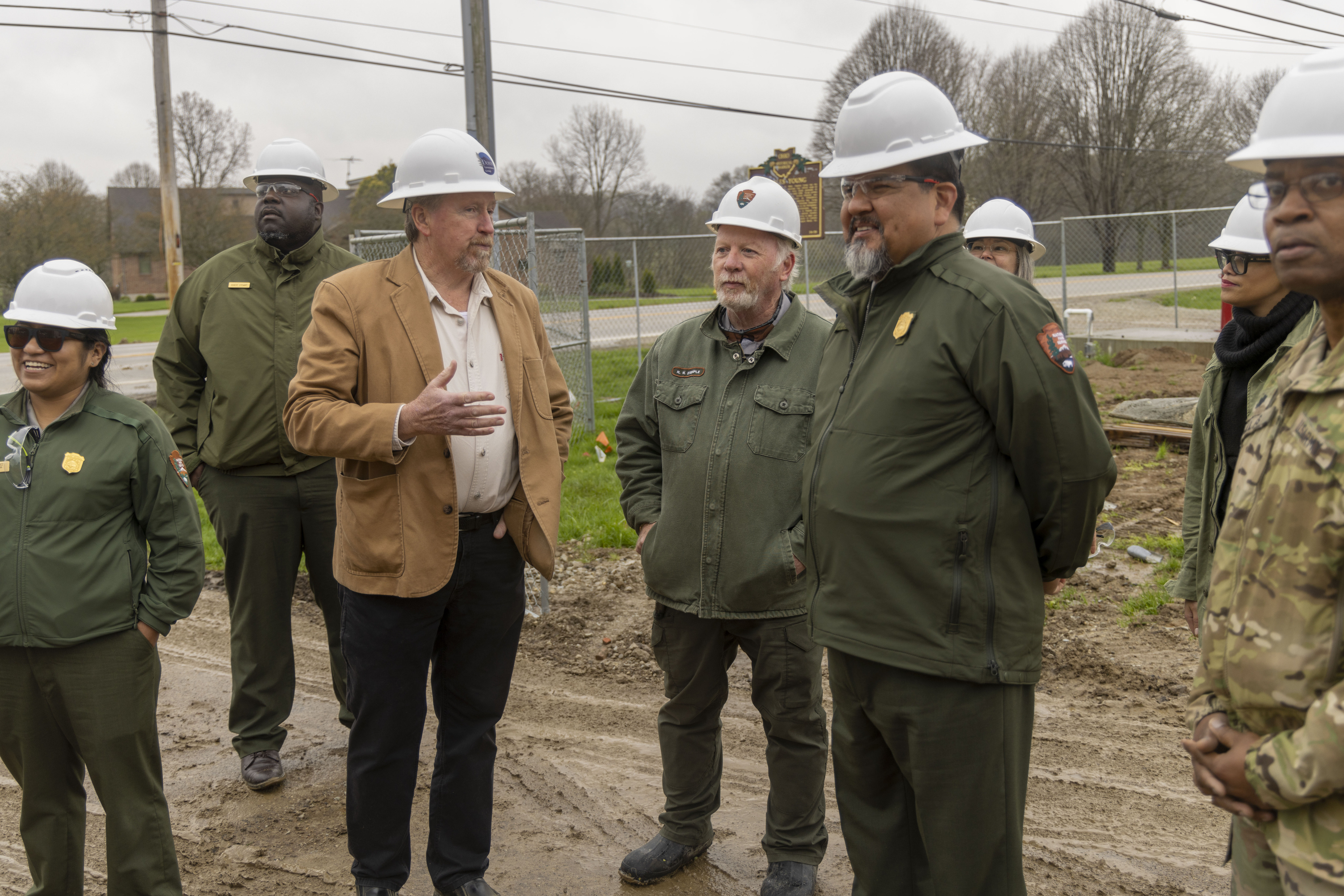 Several people wearing white hard hats listen to a man in a brown jacket speak to them all
