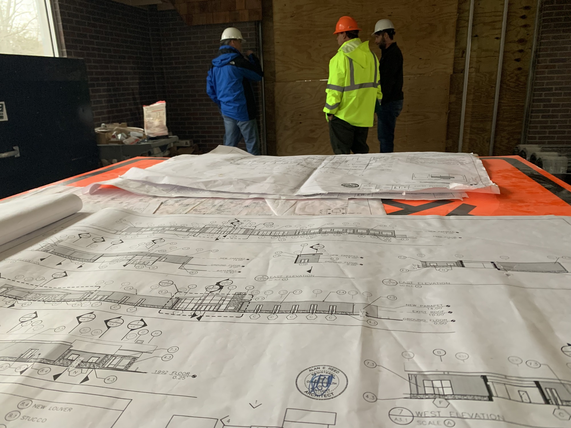 Three men talk in front of a large sheet of paper covered in architectural drawings.