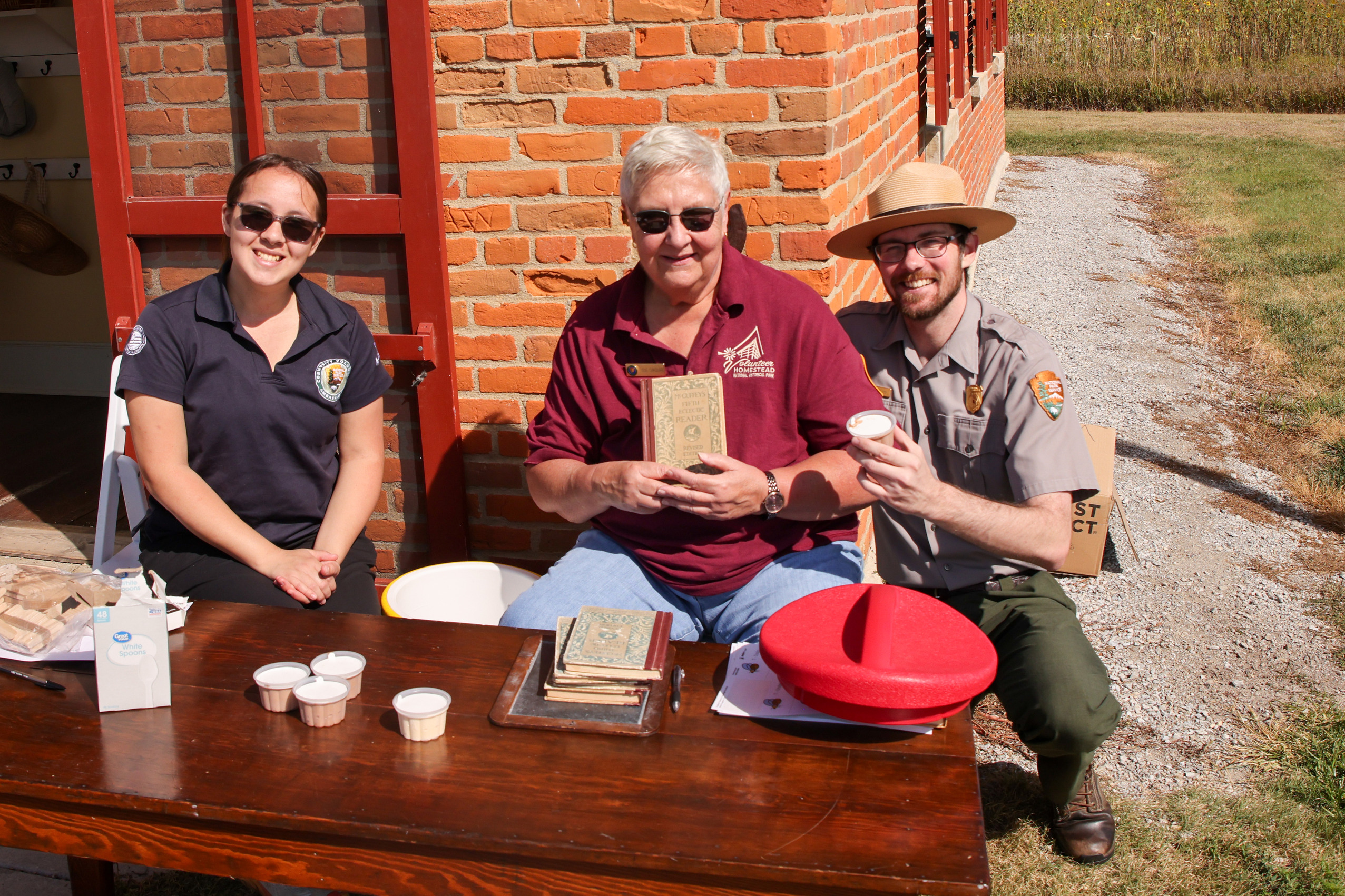 Two women in volunteer branded clothing and a uniformed ranger sit behind a table passing out small ice cream cups to visitors and contestants. The volunteer in the middle appears to be studying as she holds her "McGuffey's Fifth Eclectic Reader" up for the camera.