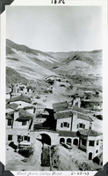 This is an historic black and white photograph from the Scotty's Castle Historic Photograph Collection, Death Valley National Park of Spanish style buildings from above. Background shows fences and buildings of an Indian Camp. Desert mountains and sky in background. Inscriptions in black ink along upper and lower border.