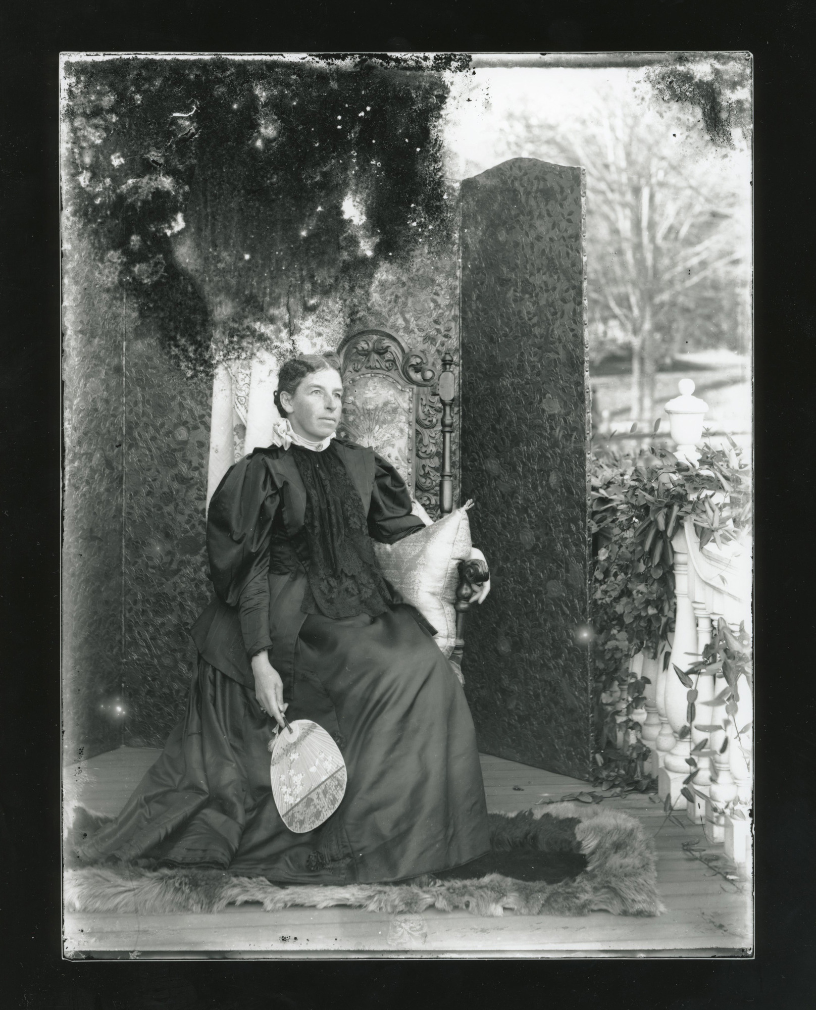 White woman in a dark-colored dress sits in an elaborate chair and holds a flat uchiwa fan down towards the ground. She sits in front of a room divider screen covered in a floral pattern. White balustrade covered in ivy at right.