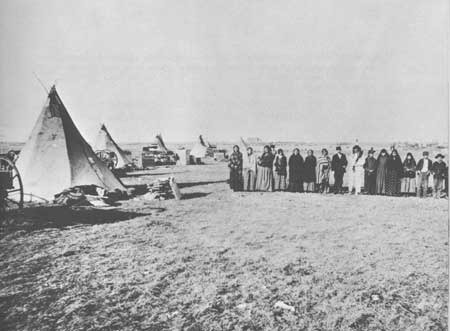 Black and white photo of an open prairie and tipis to the left with a group of people posing in a line to the right.