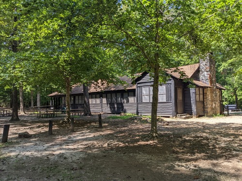 Large wooden building with a stone chimney is lined with windows and shaded by green trees