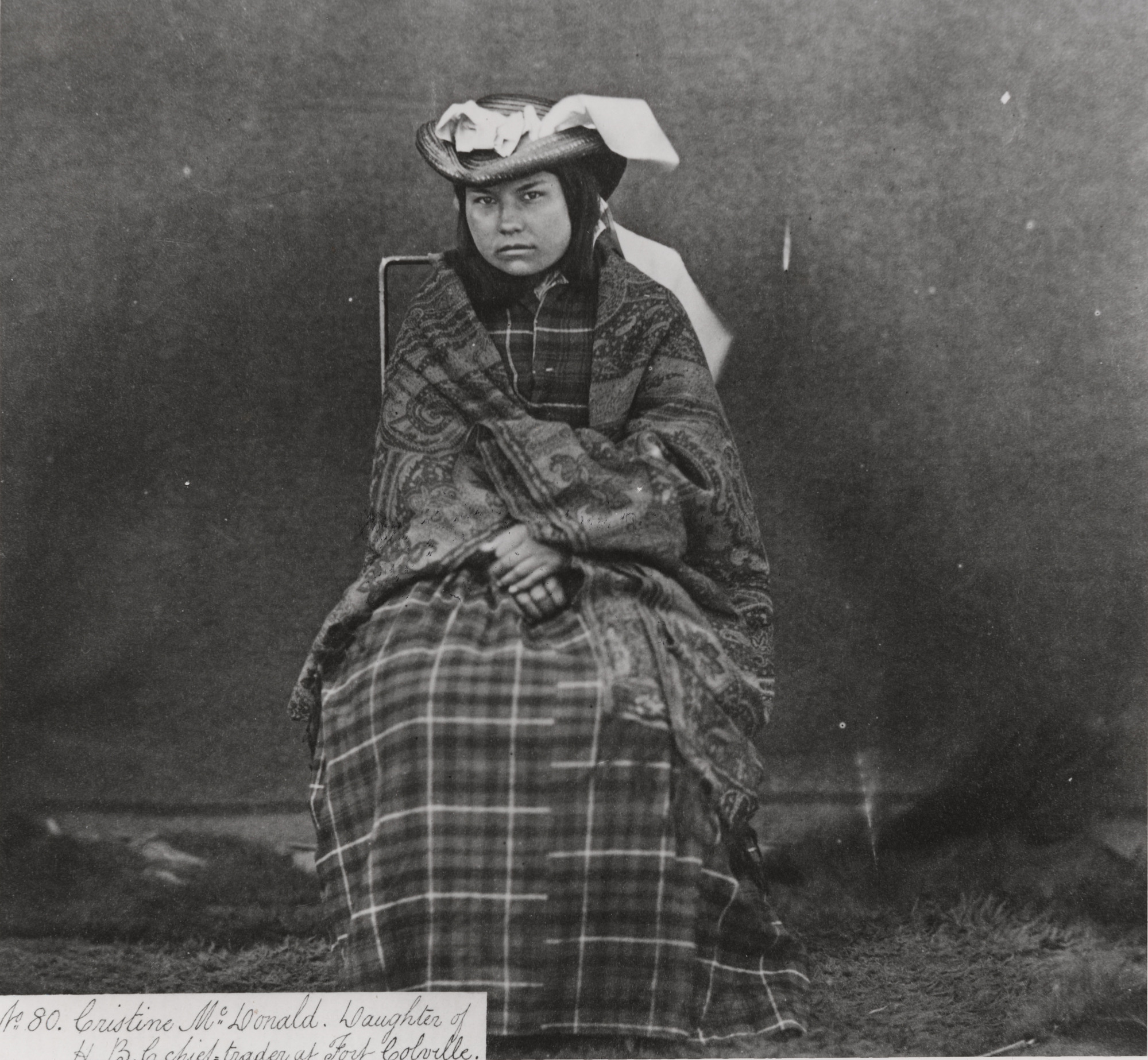 Black and white photograph of sitting woman in plaid dress, wrapped in a blanket
