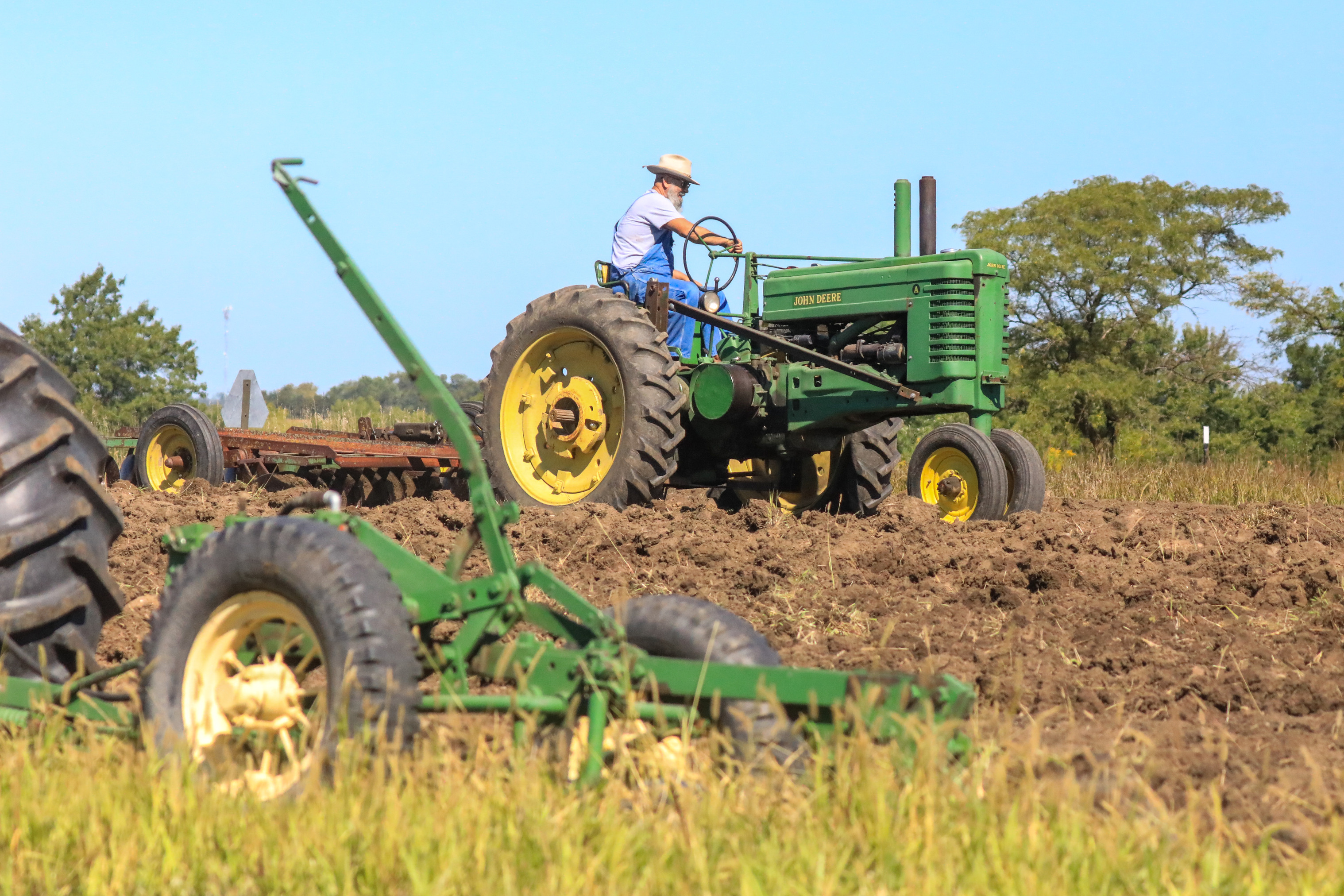 Tractor driver in straw hat and overalls turns the wheel of a green John Deere tractor on a field nearly all plowed and ready for seed.