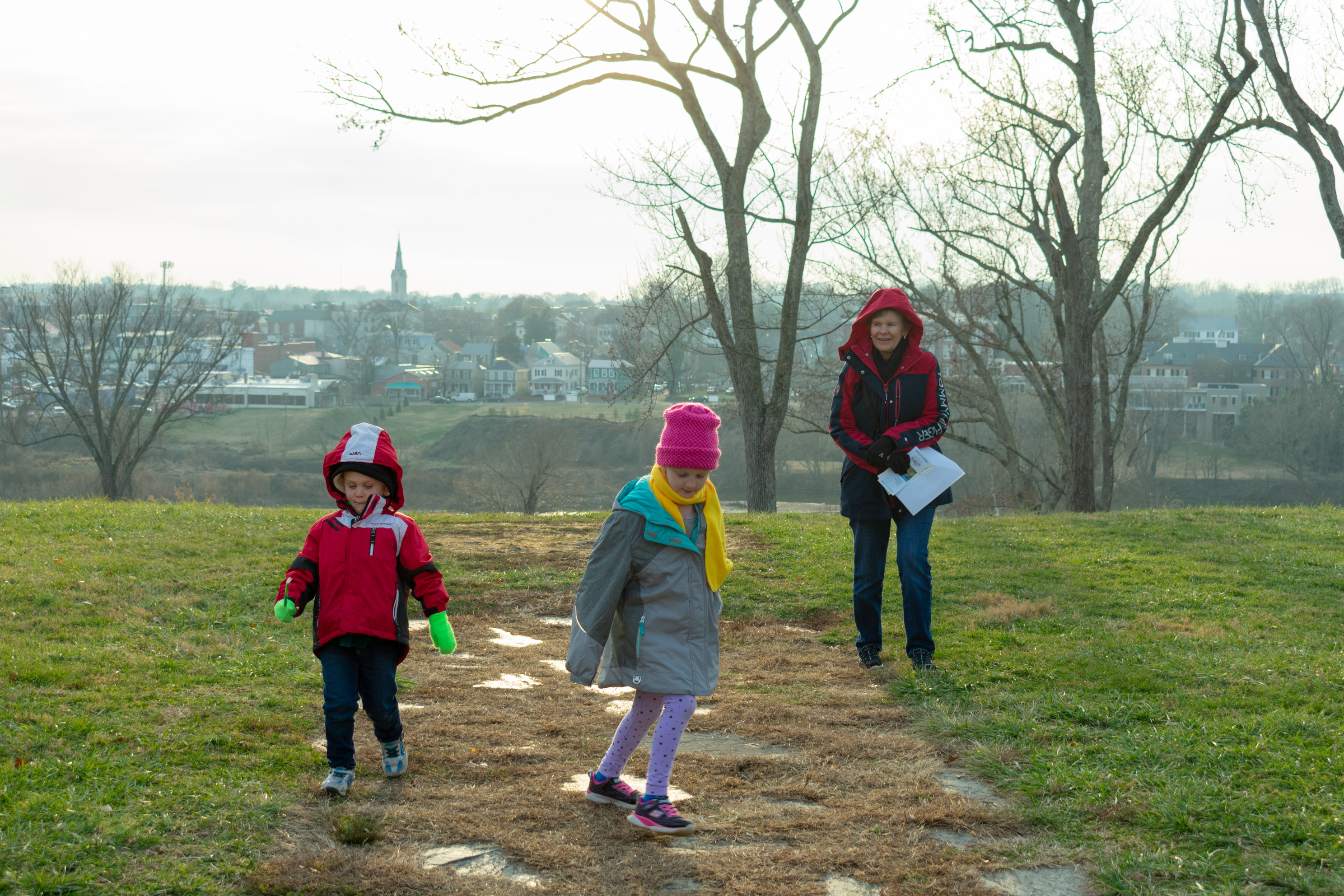 A woman and two children walk along a path overlooking a city in the distance.