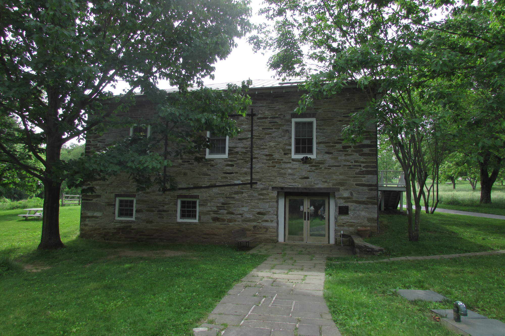 A stone walkway leads to a set of double doors in a two-story stone structure,  surrounded by grass and trees.