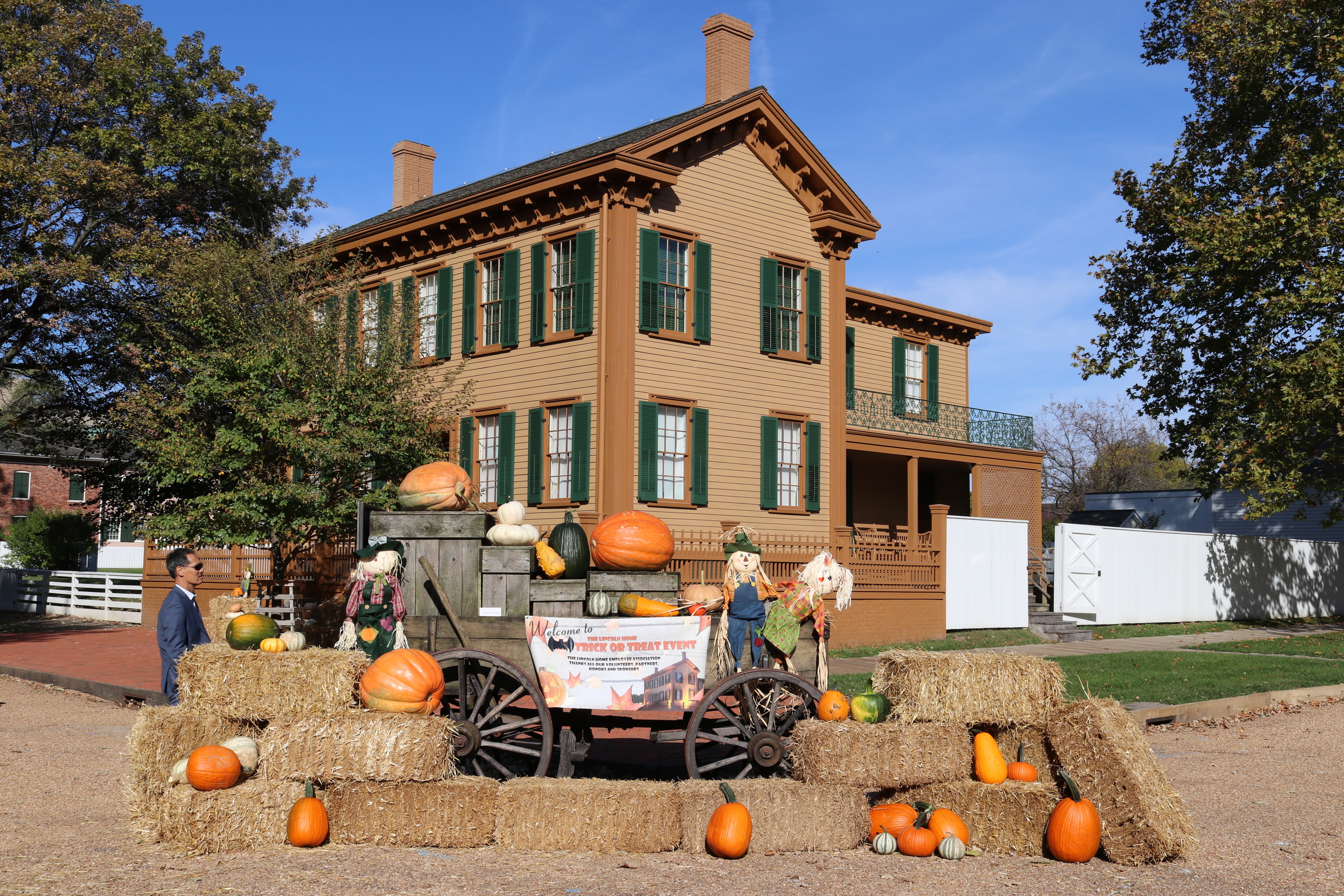 A festive fall display with hay bales, bright orange pumpkins, and a 19th century wagon sit in front of the Lincoln Home, a two-story wooden house with dark brown and green trim.