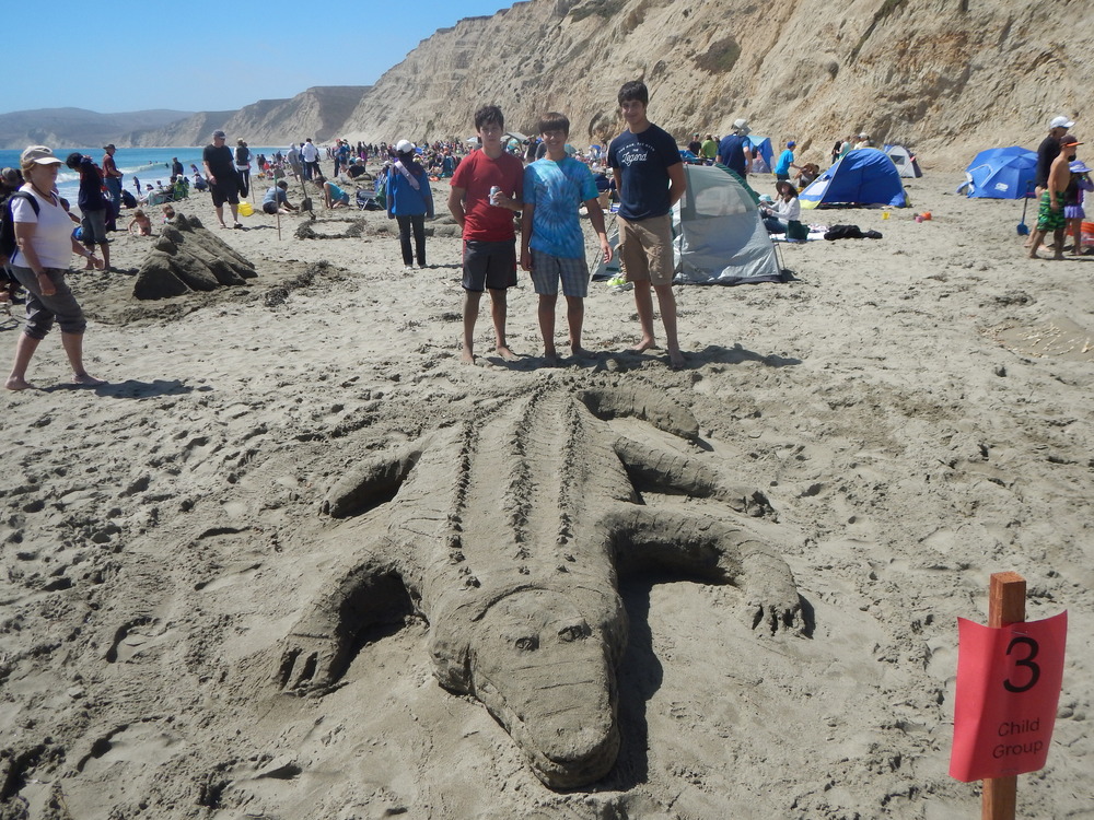 Three young boys standing behind a sand sculpture of a crocodile.