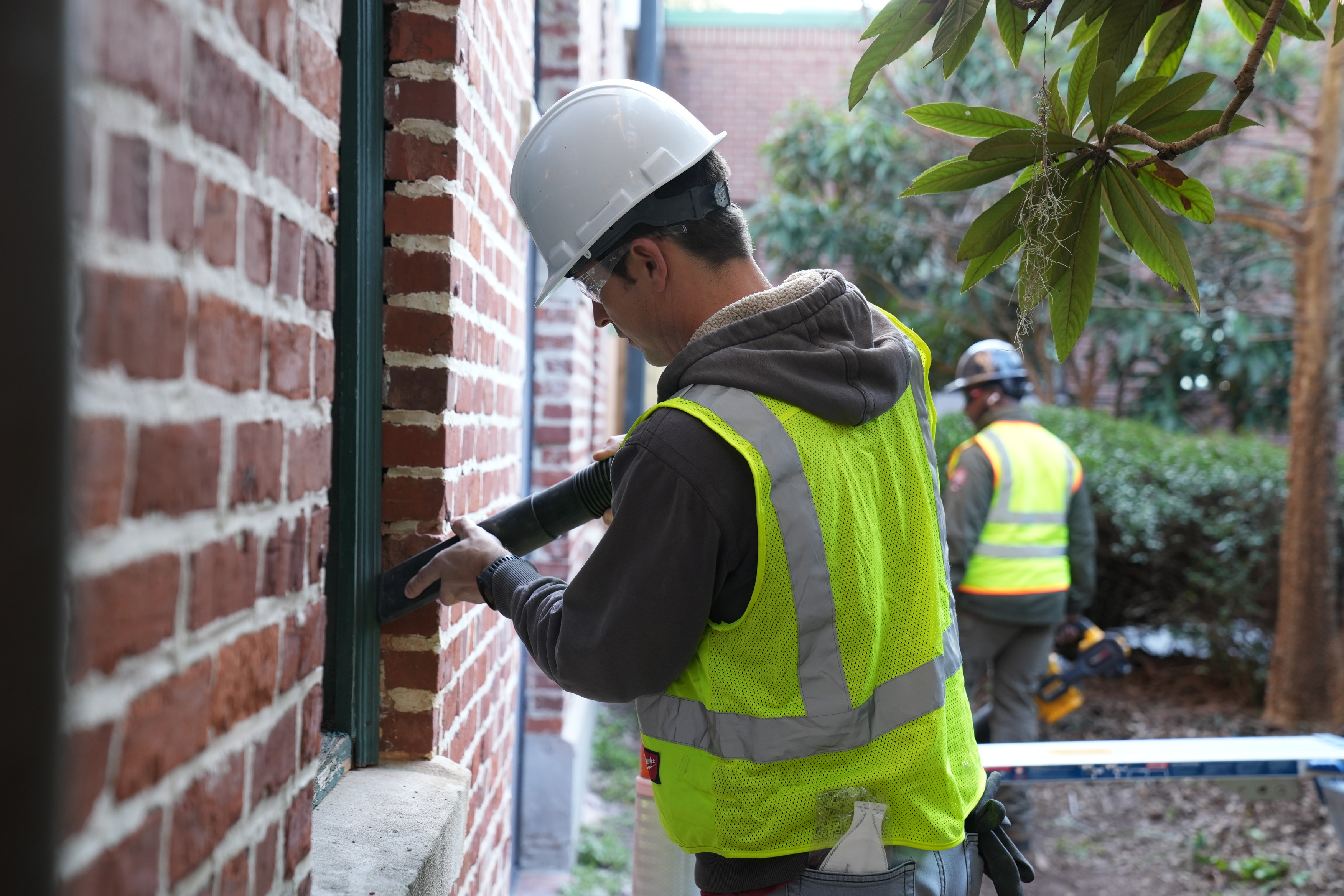 A man wearing personal protective equipment vacuums dust from a windowsill in a brick wall.