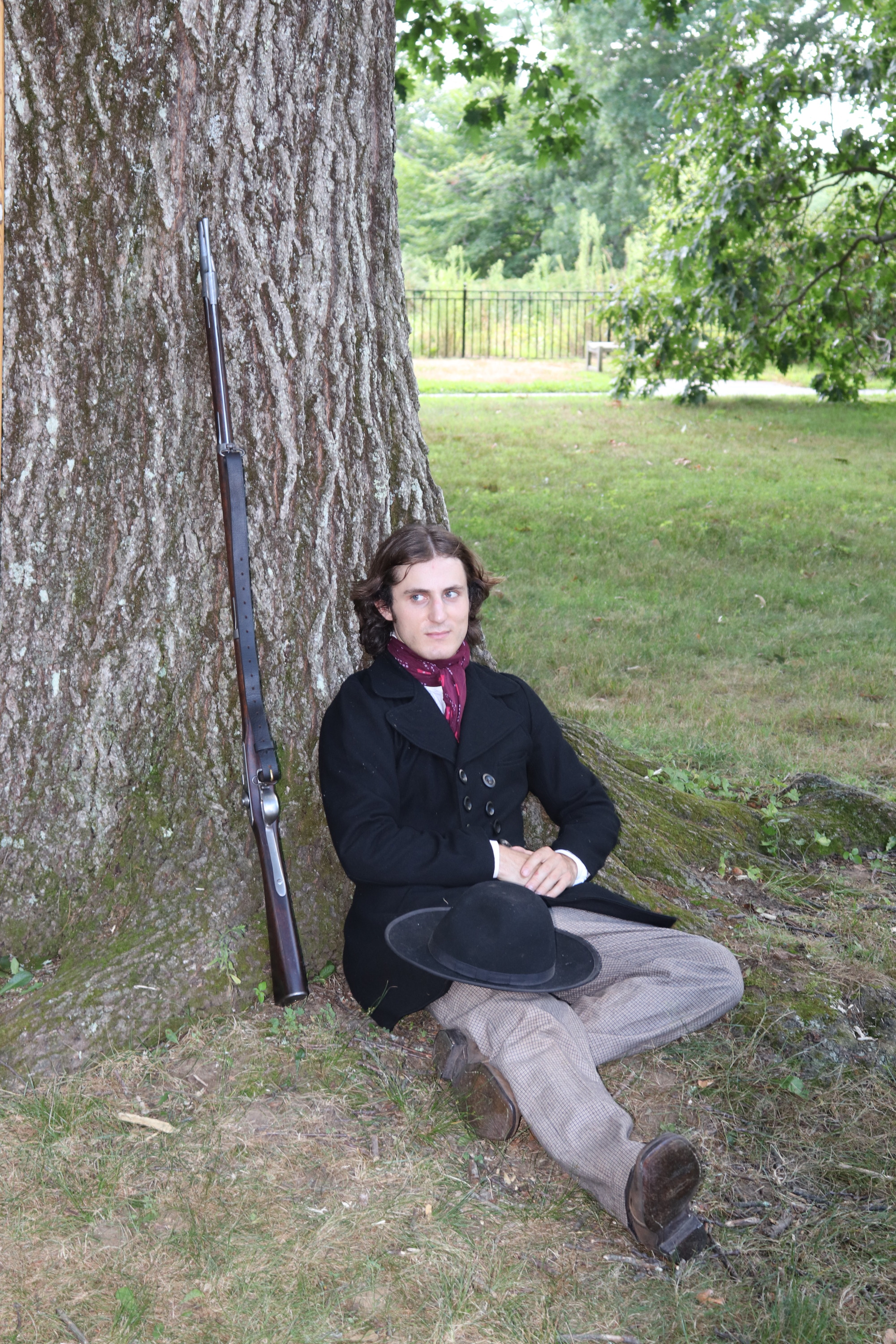 A young man, rests under a large tree, his musket leaning against the tree as well. 