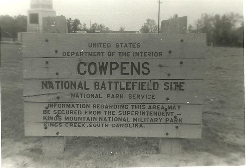 Wooden sign for Cowpens National Battlefield Site with a partial view of the US Monument behind it