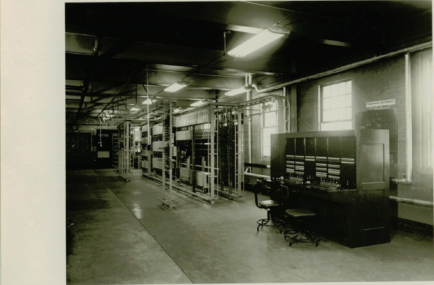 Black and white photo of a long room full of tall metal racks. A large consol desk with two chairs sits in the foreground. The metal racks either have metal boxes hanging, wires, or large panelboards. The racks are raised off the floor by being set on metal beams. Overhead lights are hung down the middle of the room.