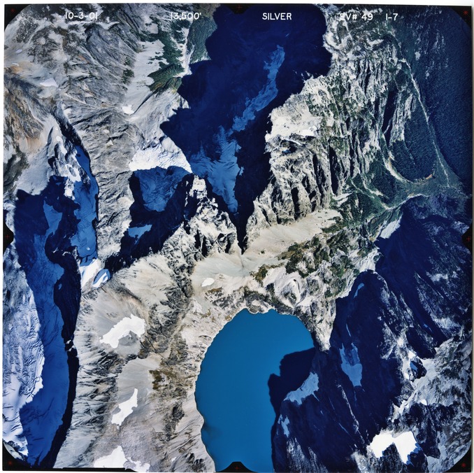 Color aerial photograph of glacier and snowfields on mountain peak with forested valleys below.