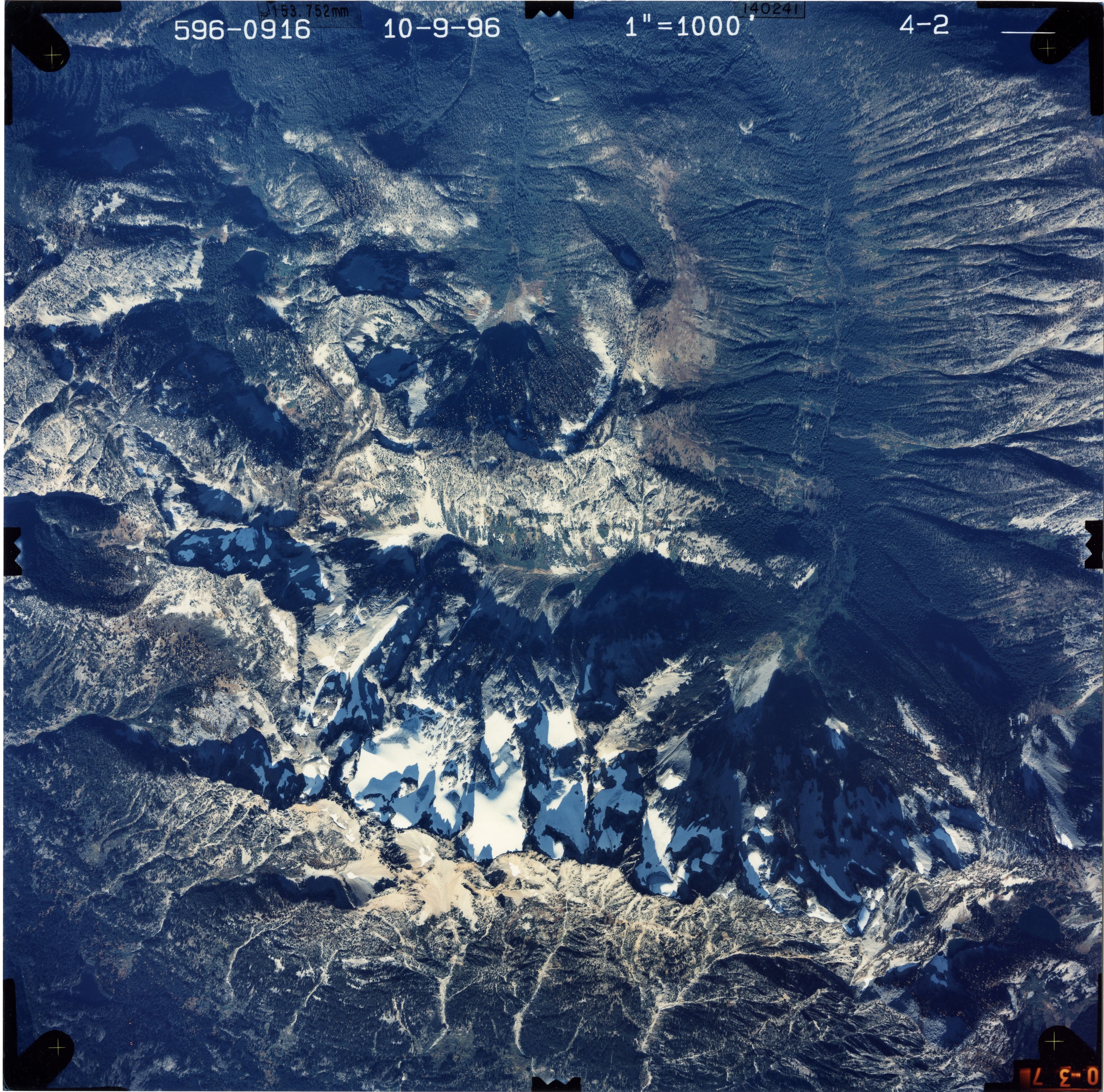 Color aerial photograph of glacier and snowfields on mountain peak with forested valleys below.