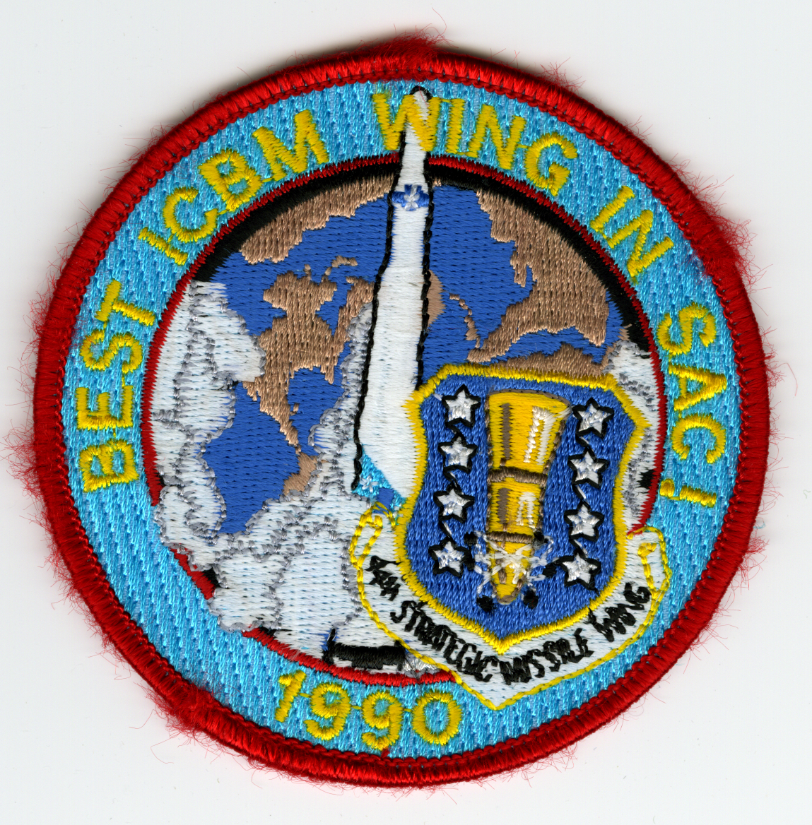 Blue circular, red border. BEST ICBM WING IN SAC! / 1990" in yellow around edge. In center is brown and blue globe with black trim and white missile with smoke coming out of bottom in center. Bottom right over globe is 44th SMW symbol."

