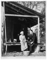 historic black and white photo of a man in a suit sitting on the porch of a woman wearing a long dress, next to a dog laying on the porch of a wooden building.