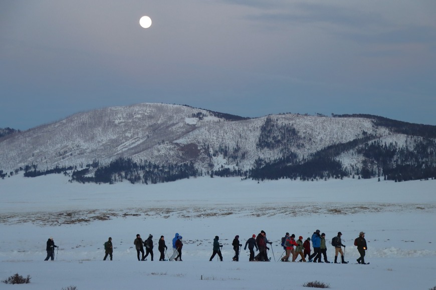 A park ranger holding a lantern guides a large group of snowshoers through a snowy valley at dusk as the full moon rises above distant mountains.