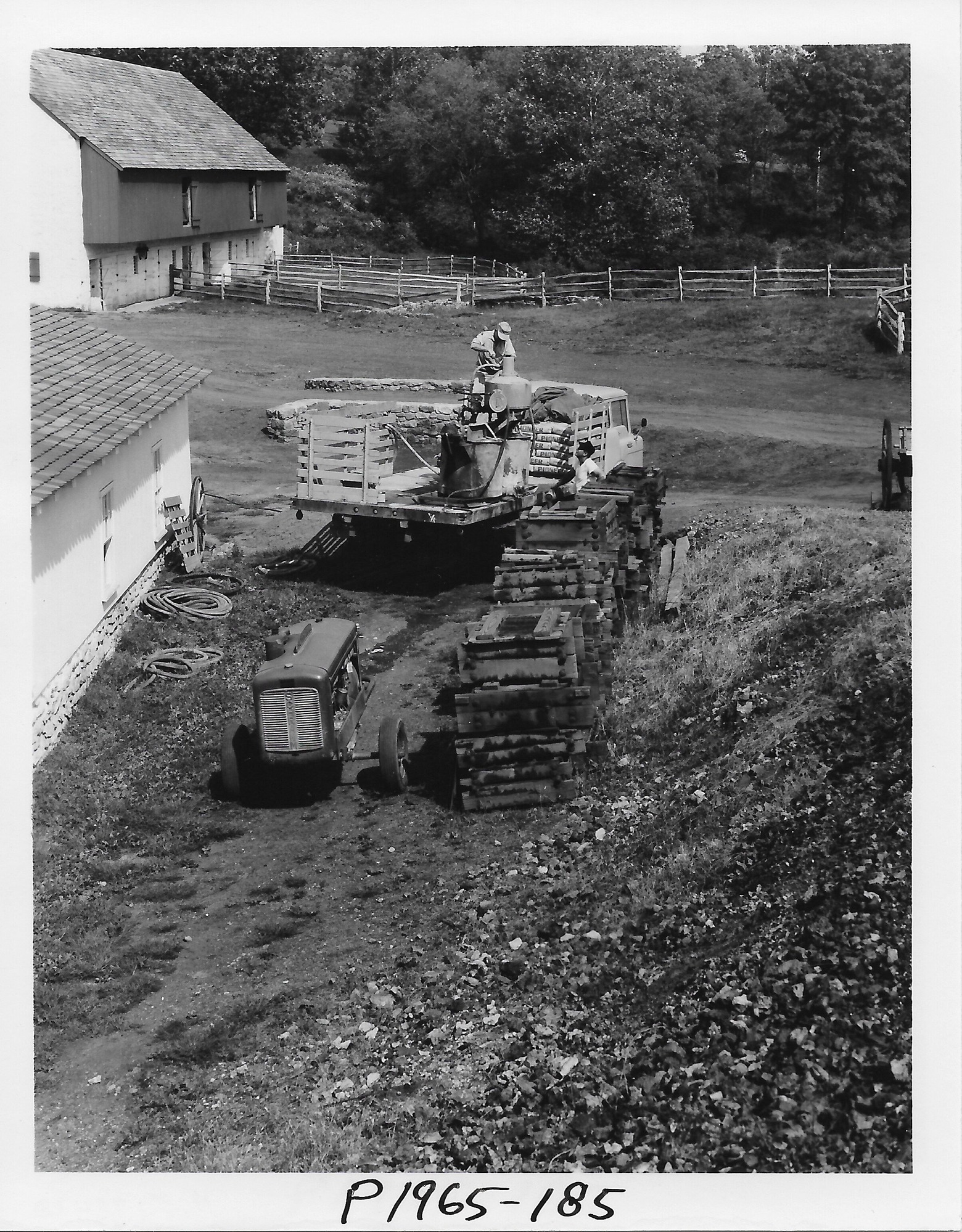 Person on top of a truck's bed working on something related to reconstruction of Cast House. Barn in the background. Tractor and wooden flasks are in the foreground. 