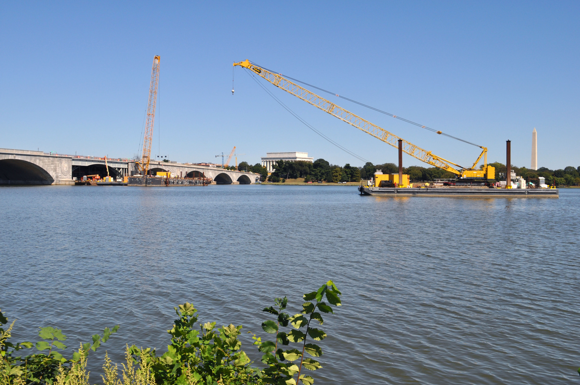 Construction on Arlington Memorial Bridge in September 2019. A crane is being driven to the bridge by a barge. The Lincoln Memorial and Washington Monument are in the background. 