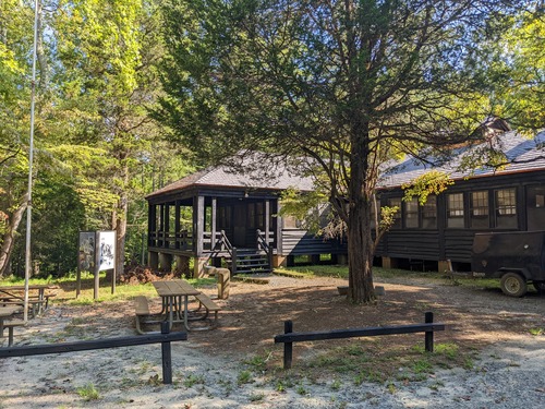 Large wooden building with windows and a porch overlooks picnic tables at the edge of a green forest