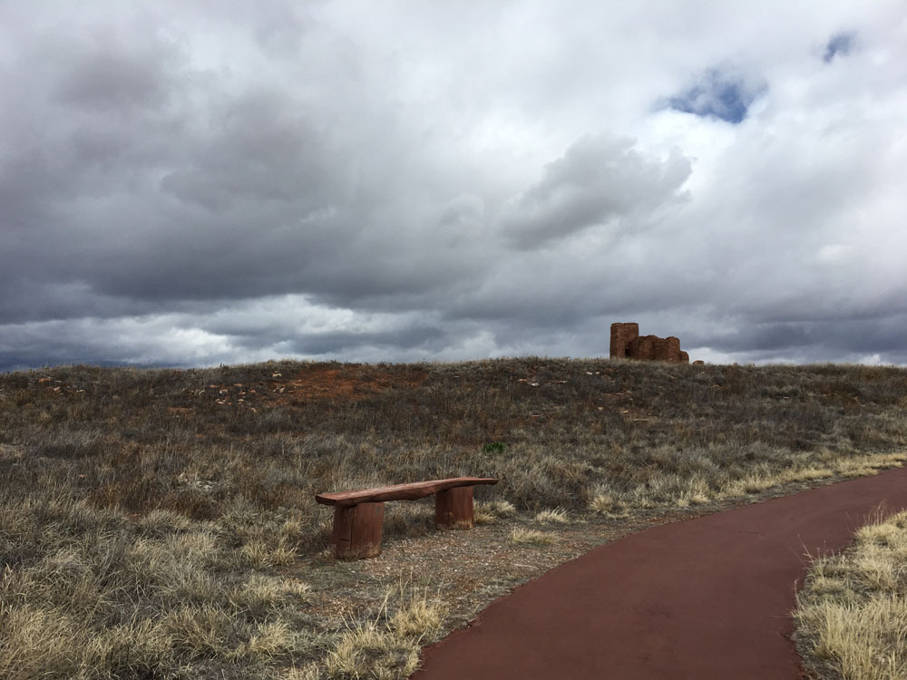 The trail winds past a wooden bench and a low hill covered in grasses with some stones scattered on it.  The remains of a stone church can be seen in the distance.