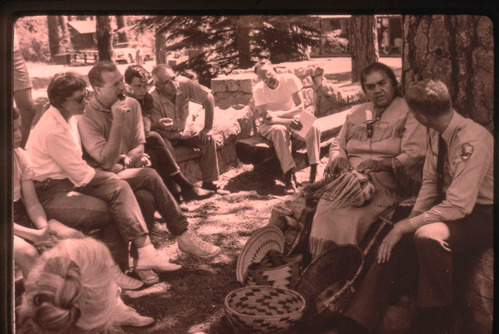 A smiling woman in American Indian attire sits with woven baskets in a demonstration. A male park ranger sits to her left.