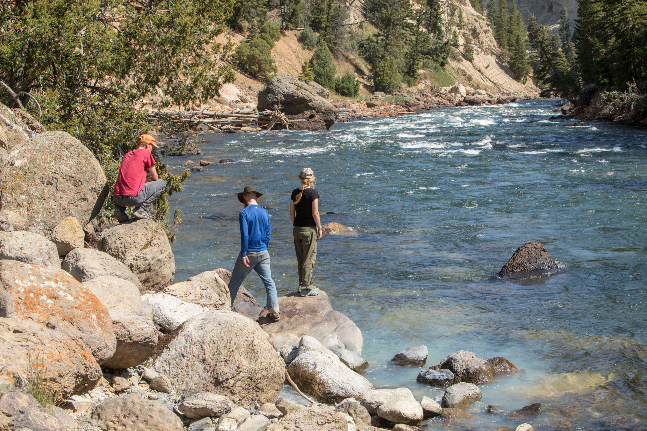 Three hikers are standing on boulders along the Yellowstone River.