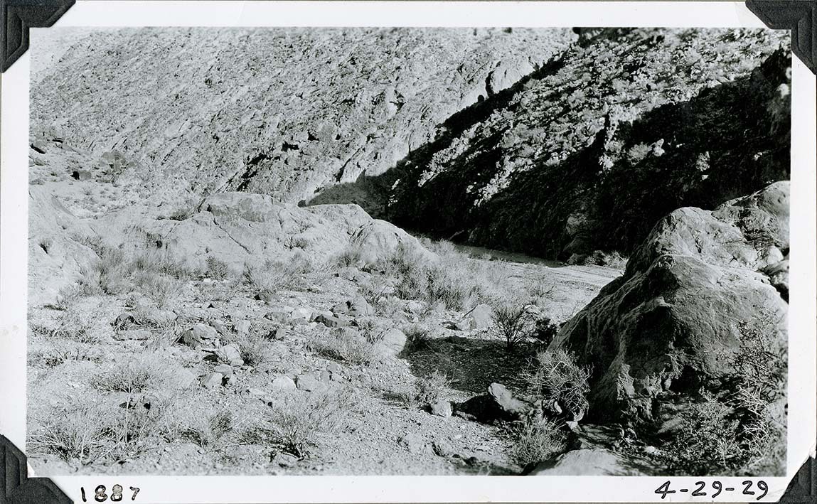This is an historic black and white photograph from the Scotty's Castle Historic Photograph Collection, Death Valley National Park of large rocks on right in foreground. Short section of dirt road visible beyond boulder. Rocky desert scene with steep canyon wall in background. Inscriptions in black ink along lower border.