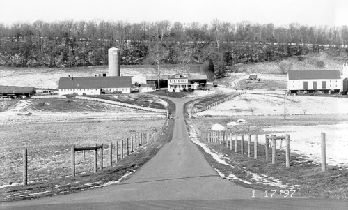 A straight, fence-lined road leads through a field to a cluster of farm buildings.