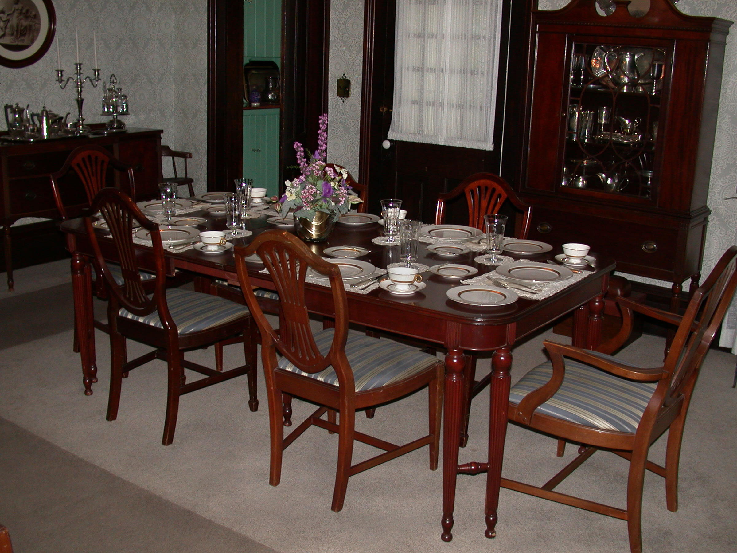 Formal dining room table, with full settings in place. Flower centerpiece. A cabinet contains several Gates/ Wallace and Truman mementos.