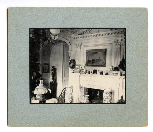 Black and white photograph of 19th century parlor, featuring small photographs on fireplace mantel.