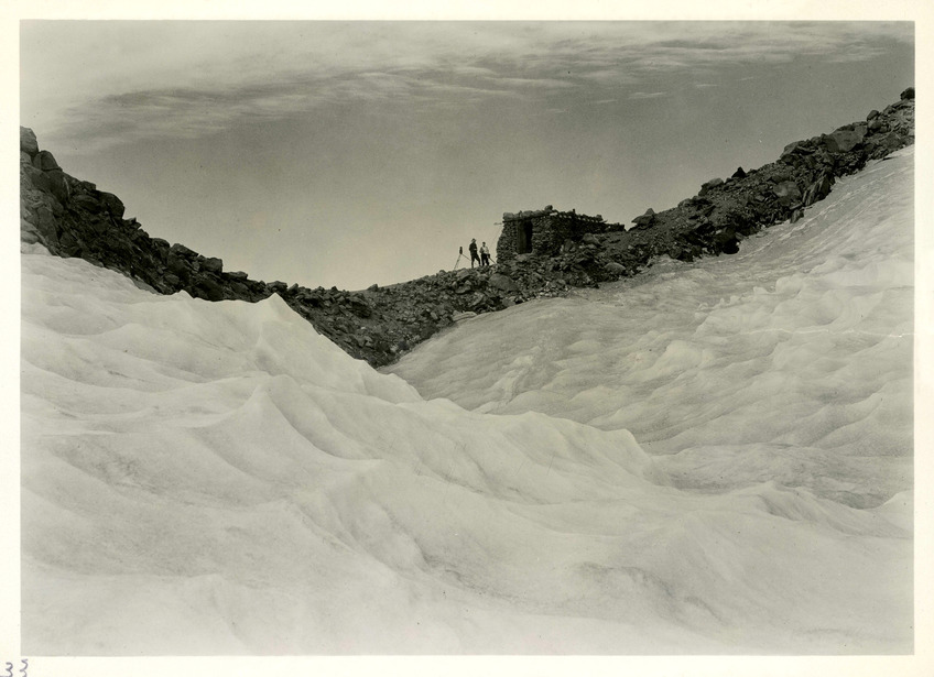 Two people stand next to a small stone building on a ridge framed by a wide sweep of glacier ice. 