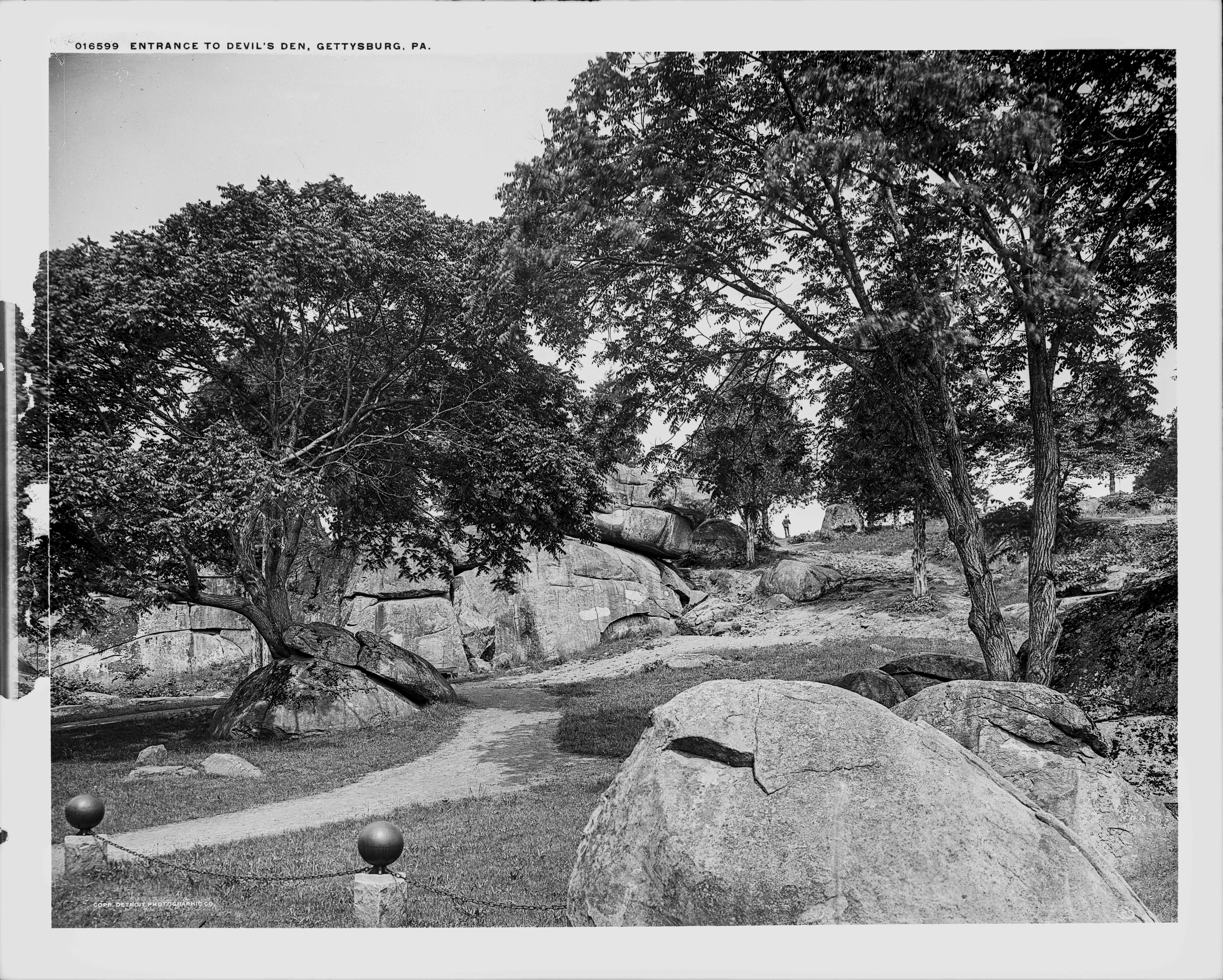 A black and white photograph with boulders, cannonball posts and chain in the foreground. A formal trail leading towards a mature tree growing next to a boulder can be seen in the middle of the picture. In the background the large boulders of Devil's Den, along with a bronze statue atop Smith’s 4th New York Artillery can be seen.