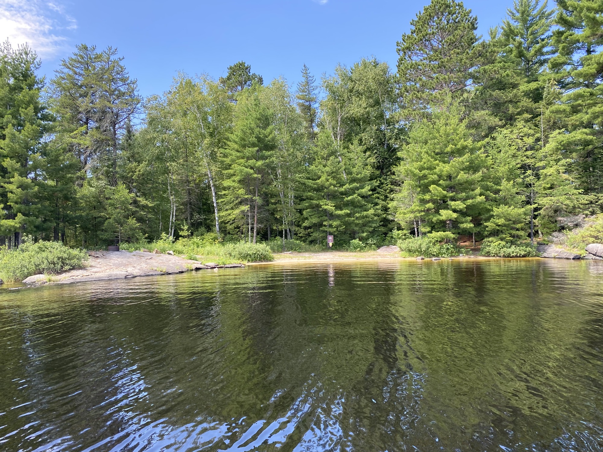 View of houseboat site from water of the beach access with the houseboat sign on edge of tree line. The campfire ring is perched on bald rock point left of image with trees covering the background.