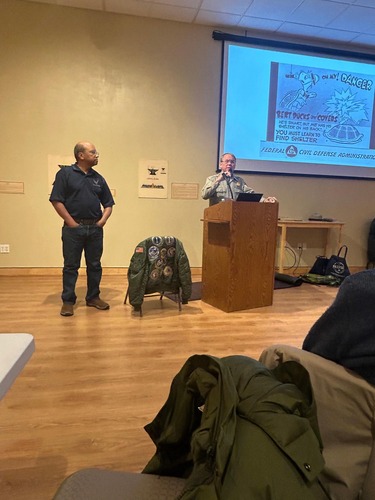Man in gray park ranger shirt standing at a podium speaking into a microphone. Man in blue shirt and blue jeans standing next to him. On the wall beside them is projected an image with a cartoon turtle.