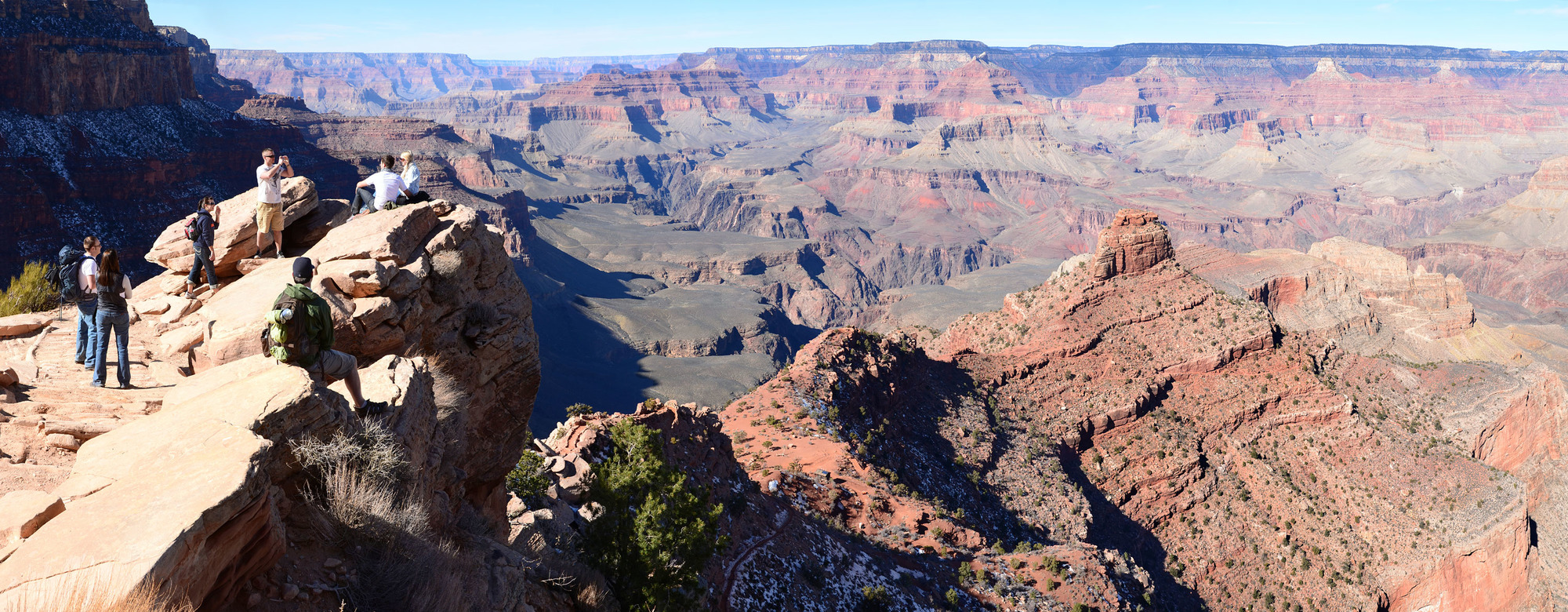 several people standing and sitting on a sloping sandstone ledge are overlooking an expansive canyon landscape