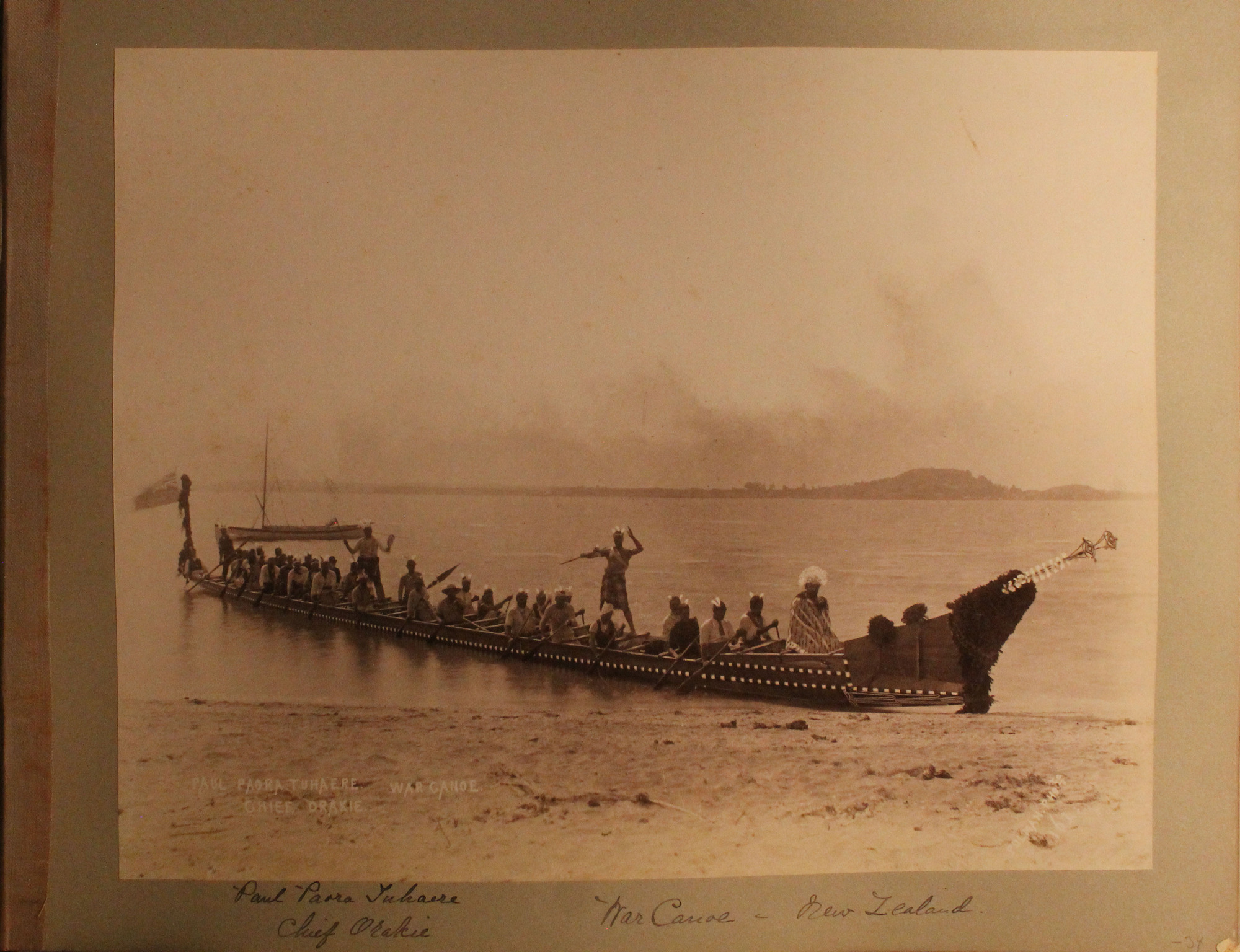 Thirty-three men sit in a long canoe next to a beach.  Distant shore in background.