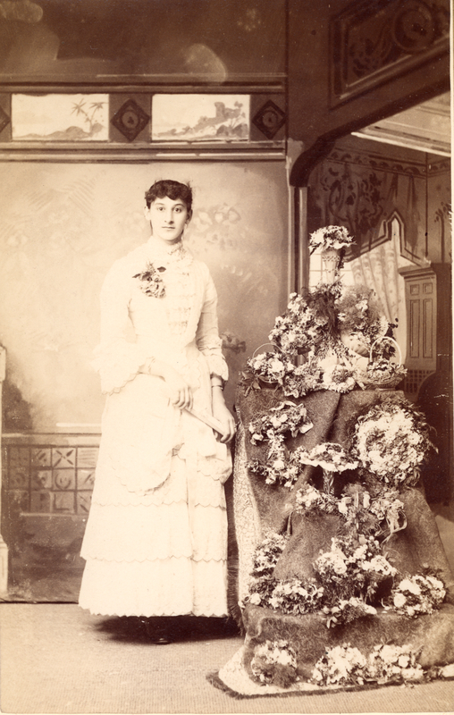 A studio portrait of Madge Gates Wallace. She is wearing a white gown, posing by flowers.