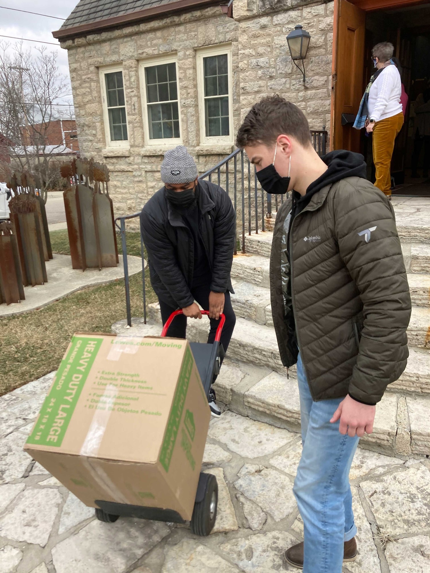 Two men in winter coats moving boxes on a hand truck, up a set of stairs into a building.